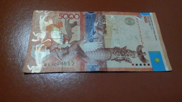 БАНКНОТА 5 000 ТЕНГЕ № серии ВК4074655 на коричневой поверхн. Одиннадцатого Января - ДЕНЕЖНЫЙ ПОТОК