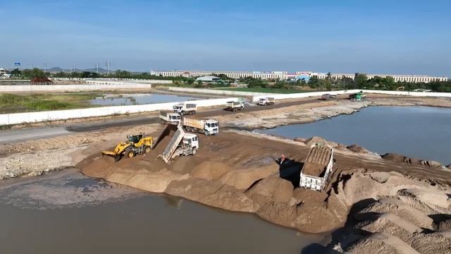 Несколько самосвалов с песком ждут, чтобы засыпать озеро песком, а колесные погрузчики толкают песок