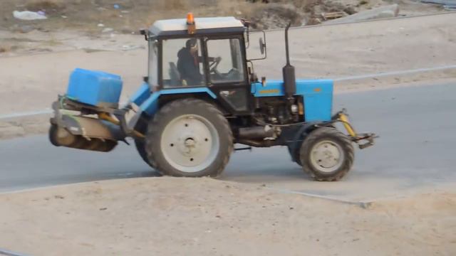 Трактор с Баком воды и Щёткой едет, чистит дорогу от грязи и пыли.