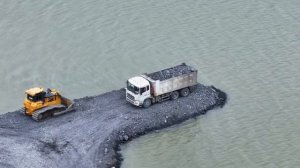 Бульдозер SHHANTUI, толкающий камни в воду, и грузовик SHACMAN.