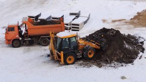 Зимой экскаватор-погрузчик загружает самосвал.