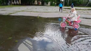 София и Давид на велосипедах катаются по лужам в городском парке.
